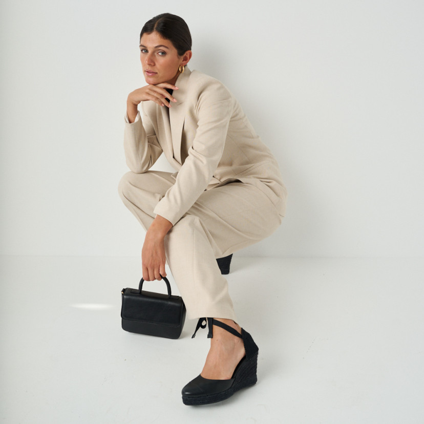 Creta leather wedge espadrille in black in full shot of the model crouching with one leg stretched out and leather bag - MONPIEL