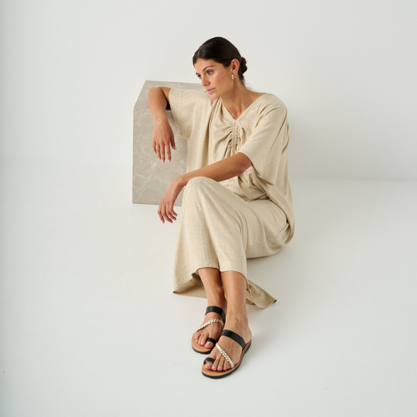 Mykonos leather Greek sandal in black, model sitting on the floor leaning on a marble cube, perspective view - MONPIEL