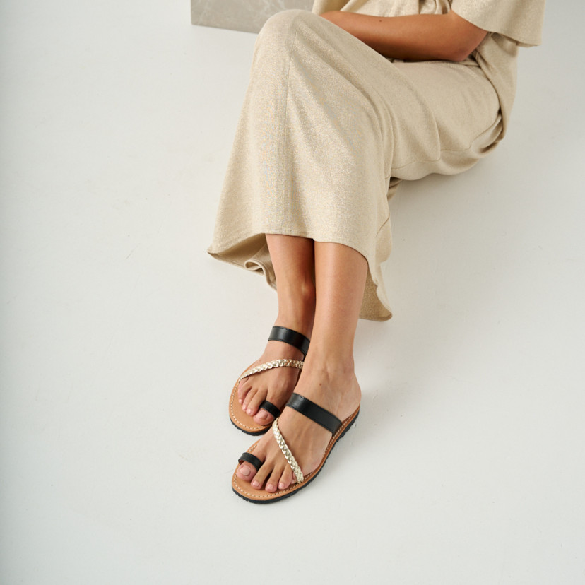 Mykonos leather Greek sandal in black, model sitting on the floor leaning on a marble cube, perspective view, medium s - MONPIEL