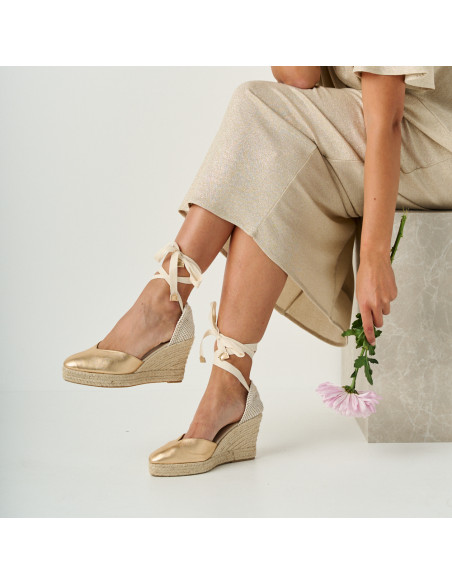 Gold leather wedge espadrille Elba with side view, crossed legs, flower detail in hand - MONPIEL Gold leather wedge espadrille Elba with side view, crossed legs, flower detail in hand - MONPIEL