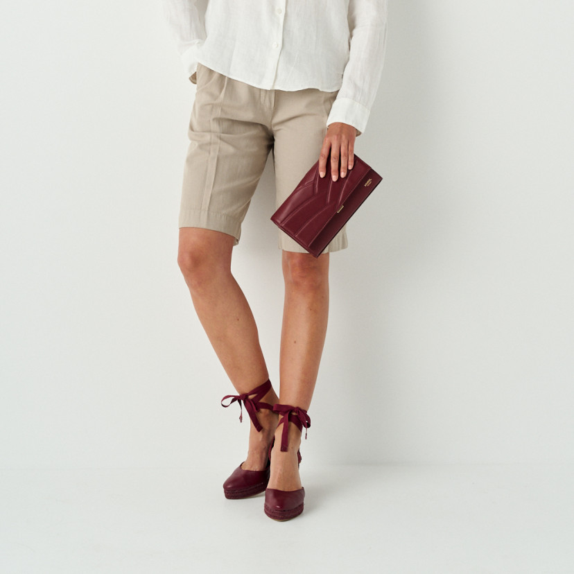 Venice women’s leather clutch bag in burgundy held in hand with detail of burgundy wedges - MONPIEL