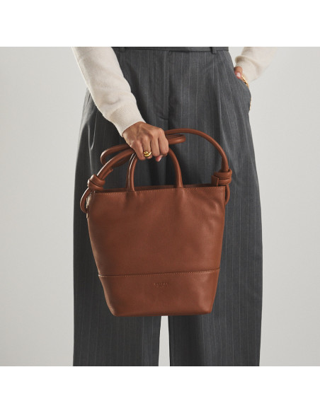 Large leather knot handbag in leather color with front view, held by the short handle in the hand - MONPIEL Large leather knot handbag in leather color with front view, held by the short handle in the hand - MONPIEL
