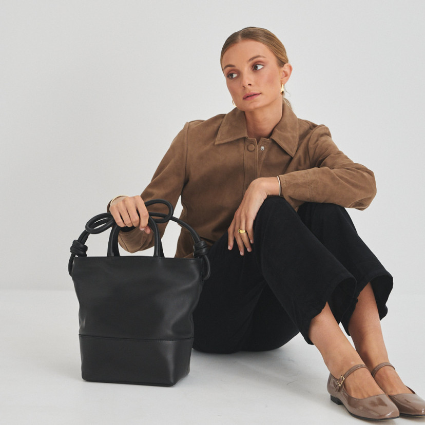 Bolsa grande com nós de couro na cor preta com modelo sentada no chão, vista frontal da bolsa e plano geral - MONPIEL