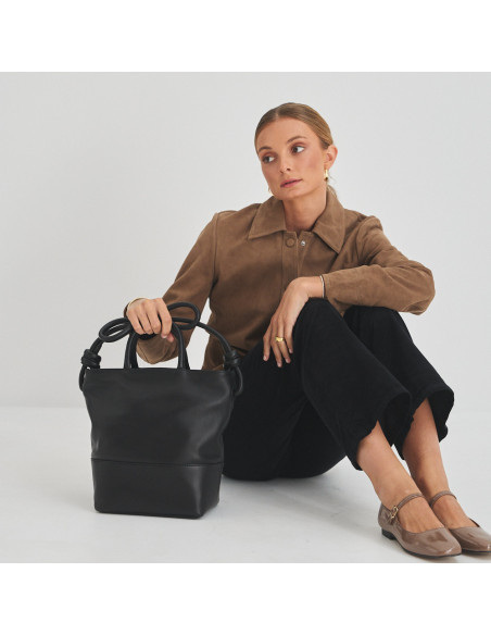 Bolsa grande com nós de couro na cor preta com modelo sentada no chão, vista frontal da bolsa e plano geral - MONPIEL Bolsa grande com nós de couro na cor preta com modelo sentada no chão, vista frontal da bolsa e plano geral - MONPIEL
