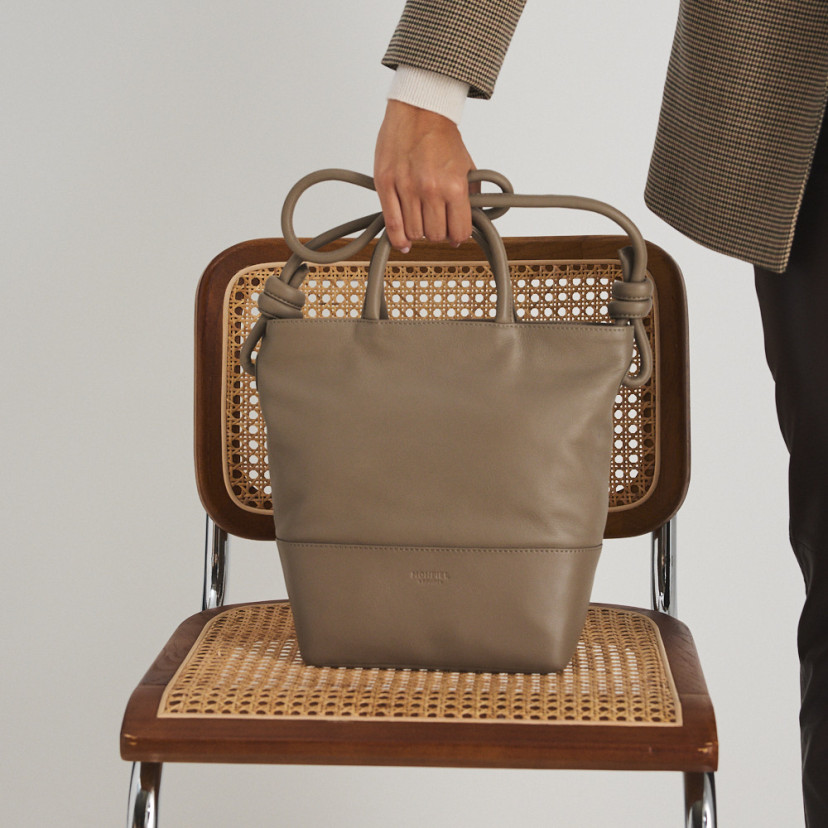 Large taupe leather knot handbag with front view, resting on a chair, and the model holding the short handle - MONPIEL