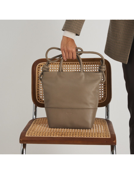 Bolsa grande com nós de couro na cor taupe com vista frontal, apoiada sobre uma cadeira, modelo segurando a alça curta - MONPIEL