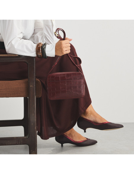 Bolsa a tiracolo de couro bordô com estampa de crocodilo segurada pela alça longa com modelo sentado - MONPIEL Bolsa a tiracolo de couro bordô com estampa de crocodilo segurada pela alça longa com modelo sentado - MONPIEL