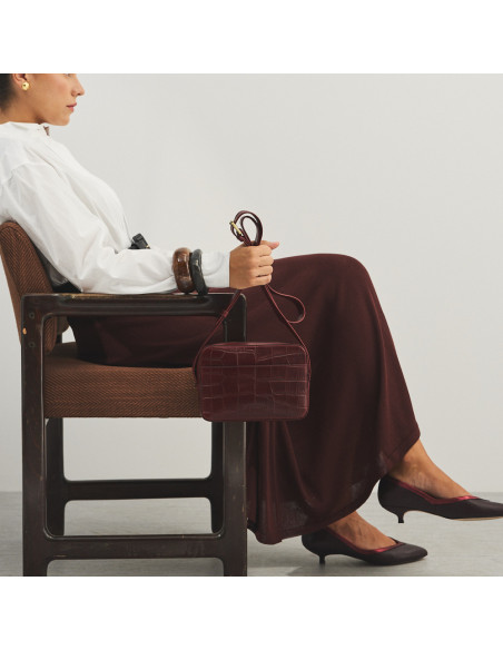Sac bandoulière en cuir bordeaux avec imprimé crocodile, tenu par l’anse, modèle assis de profil - MONPIEL Sac bandoulière en cuir bordeaux avec imprimé crocodile, tenu par l’anse, modèle assis de profil - MONPIEL