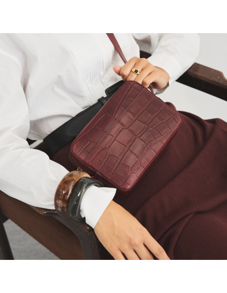 Bolsa a tiracolo de couro bordô com estampa de crocodilo usada como bandoleira com modelo sentada na cadeira - MONPIEL Bolsa a tiracolo de couro bordô com estampa de crocodilo usada como bandoleira com modelo sentada na cadeira - MONPIEL