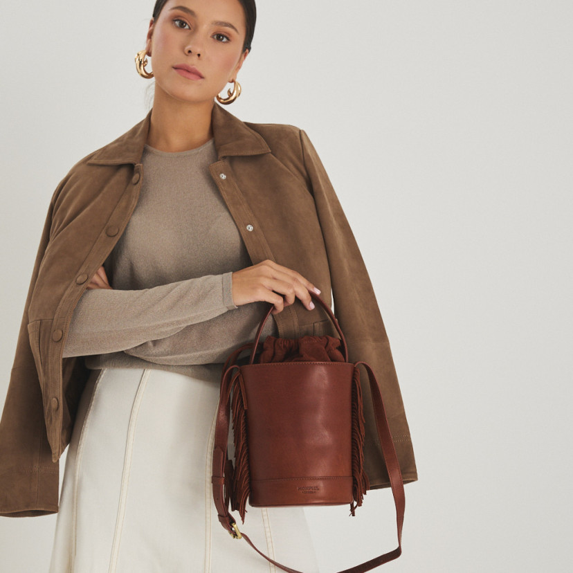Bolsa bucket de couro com franjas pequena Roma na cor couro, vista frontal, segurada pela alça curta, com jaqueta de c - MONPIEL