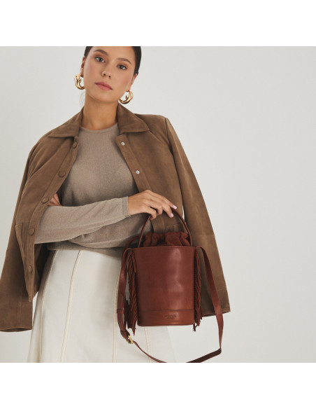 Bolsa bucket de couro com franjas pequena Roma na cor couro, vista frontal, segurada pela alça curta, com jaqueta de c - MONPIEL