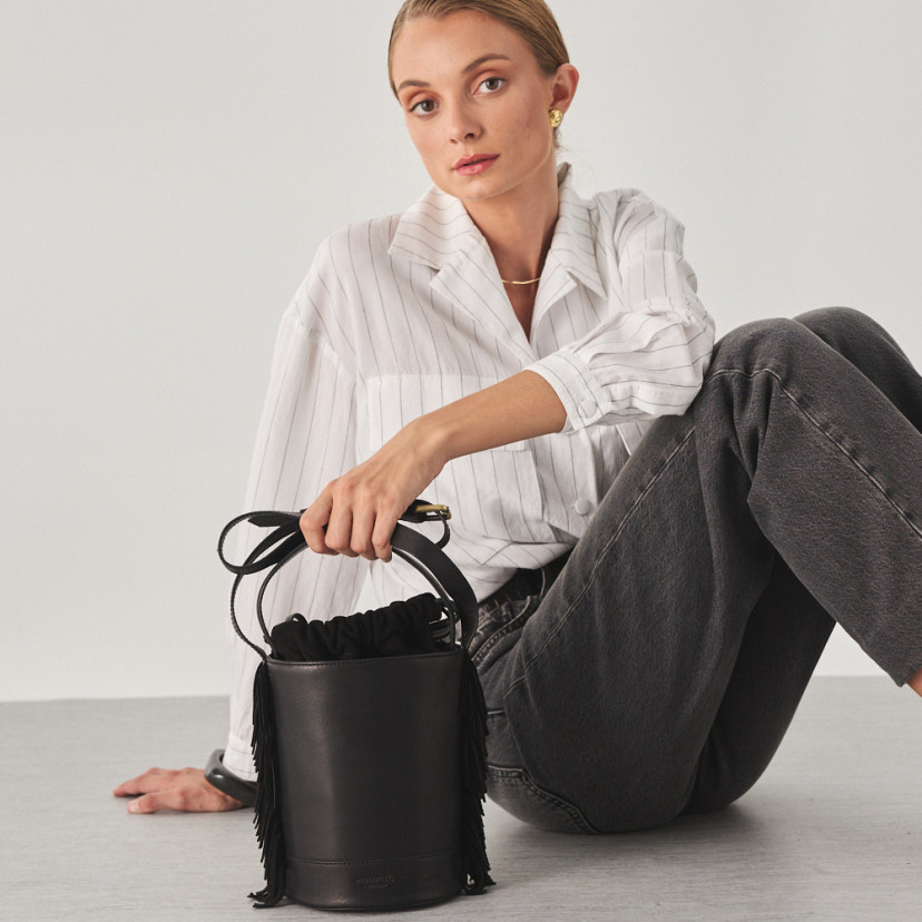 Small Roma leather fringe bucket bag in black, front view, resting on the floor - MONPIEL