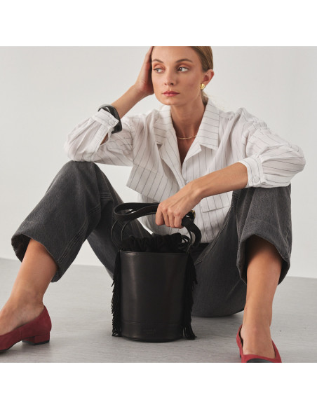 Bolsa bucket de couro com franjas pequena Roma na cor preta, vista frontal, apoiada no chão com modelo sentado no chão - MONPIEL