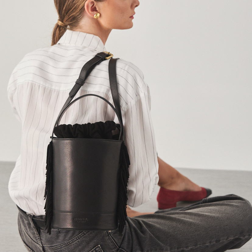 Bolsa bucket de couro com franjas pequena Roma na cor preta, pendurada nas costas com modelo sentado no chão - MONPIEL