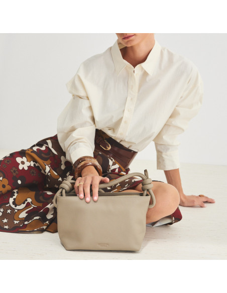 Bolsa de couro com nós pequena na cor taupe, apoiada no chão, vista frontal e plano geral de modelo sentada no chão - MONPIEL