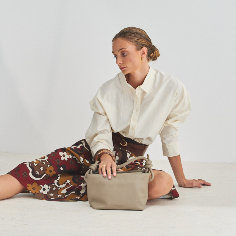 Bolsa de couro com nós pequena na cor taupe com modelo sentada no chão e mão apoiada sobre a bolsa, vista geral - MONPIEL