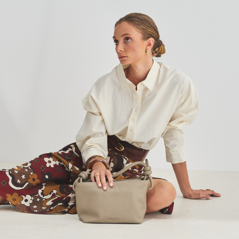 Bolsa de couro com nós pequena na cor taupe com modelo sentada no chão e mão apoiada sobre a bolsa, vista geral - MONPIEL
