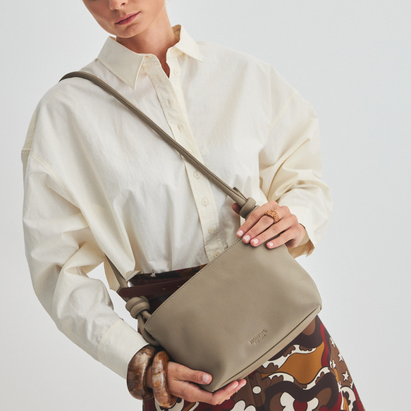 Bolsa de couro com nós pequena na cor taupe pendurada no ombro e segurada com as mãos, vista frontal e inclinada da bo - MONPIEL
