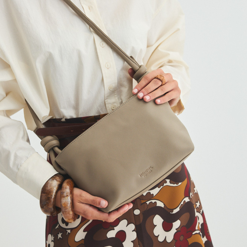 Bolsa de couro com nós pequena na cor taupe pendurada no ombro e segurada com as mãos, vista frontal e inclinada da bo - MONPIEL