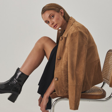 Camel leather jacket with stitching details, draped over shoulders, profile view, wide shot of model sitting on a chai - MONPIEL 2