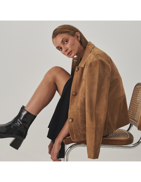 Camel leather jacket with stitching details, draped over shoulders, profile view, wide shot of model sitting on a chai - MONPIEL Camel leather jacket with stitching details, draped over shoulders, profile view, wide shot of model sitting on a chai - MONPIEL