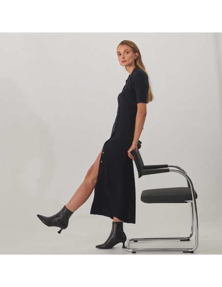 Jimena black leather pointed-toe ankle boot, profile view, wide shot of model standing, leaning on a chair with one le - MONPIEL Jimena black leather pointed-toe ankle boot, profile view, wide shot of model standing, leaning on a chair with one le - MONPIEL