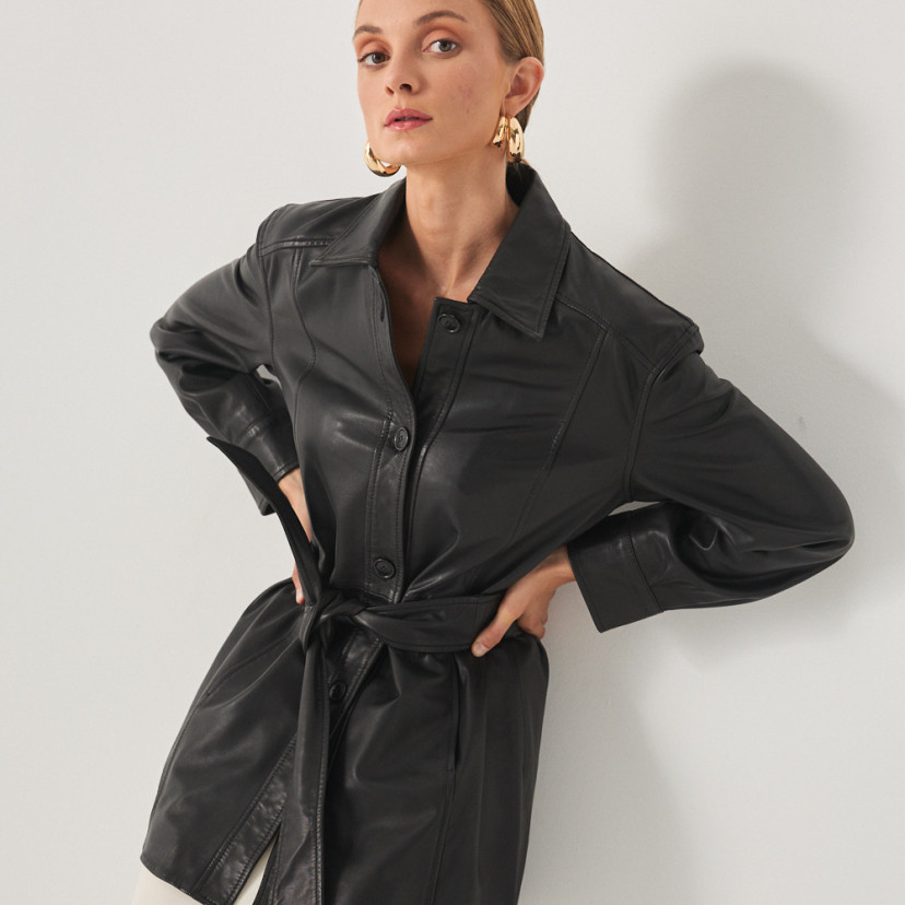 Trench de couro preto, modelo apoiada na parede com as mãos na cintura - MONPIEL