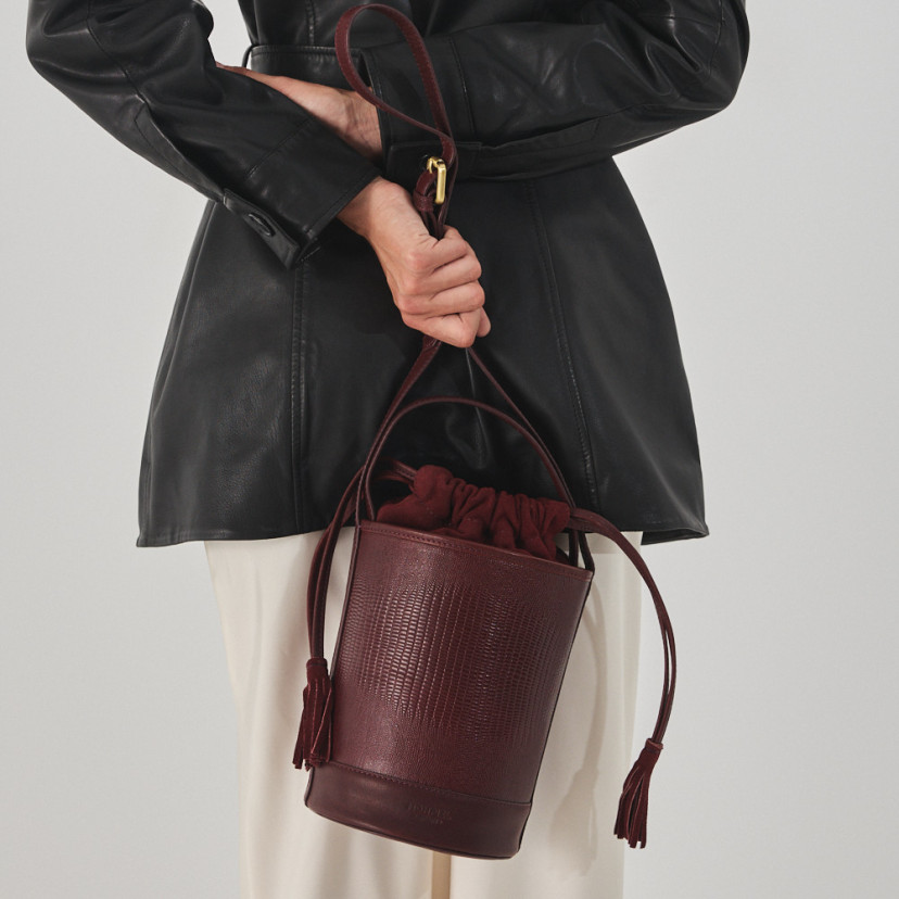 Small round bucket leather bag in burgundy snake print, front view, held at the back - MONPIEL