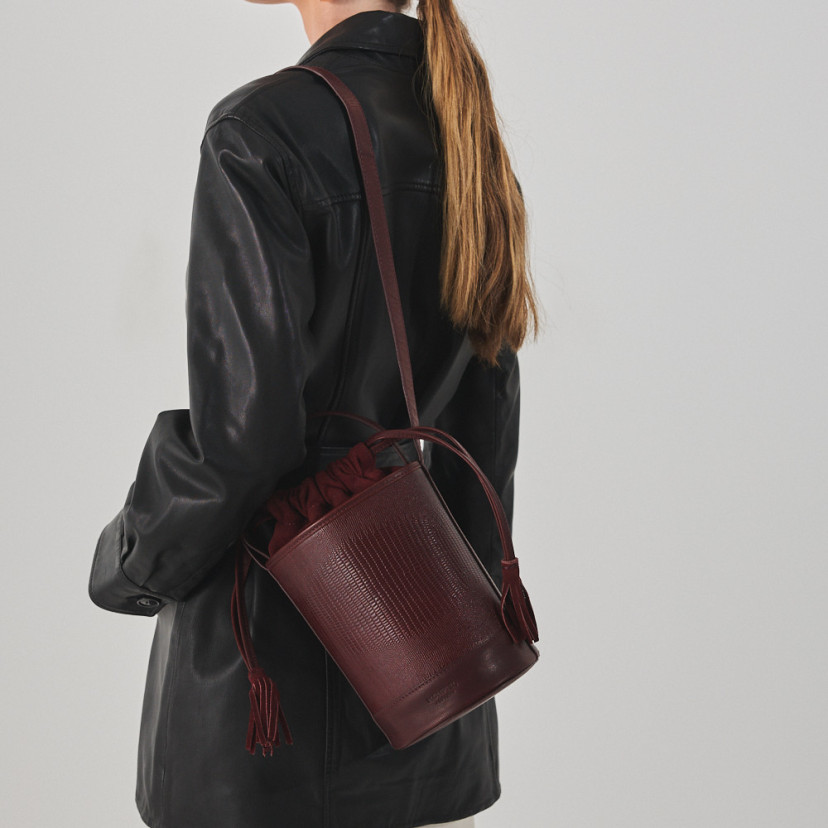 Bolsa bucket redonda pequena de couro em cor de serpente bordô, pendurada no ombro, plano médio - MONPIEL
