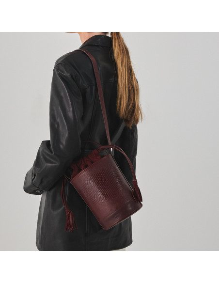 Bolsa bucket redonda pequena de couro em cor de serpente bordô, pendurada no ombro, plano médio - MONPIEL