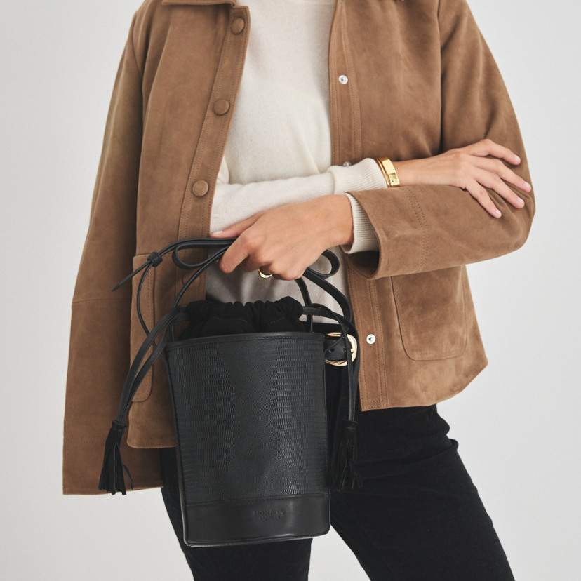 Bolsa bucket redonda pequena de couro em cor de serpente preta, vista frontal, segurada pela alça - MONPIEL