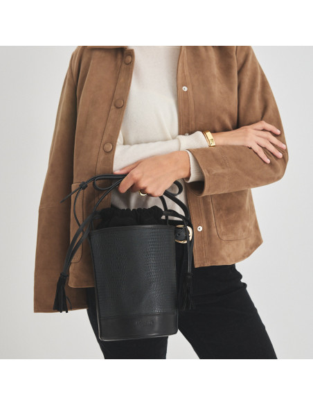 Bolsa bucket redonda pequena de couro em cor de serpente preta, vista frontal, segurada pela alça - MONPIEL