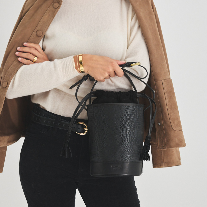 Bolsa bucket redonda pequena de couro em cor de serpente preta, vista frontal, segurada pela alça - MONPIEL
