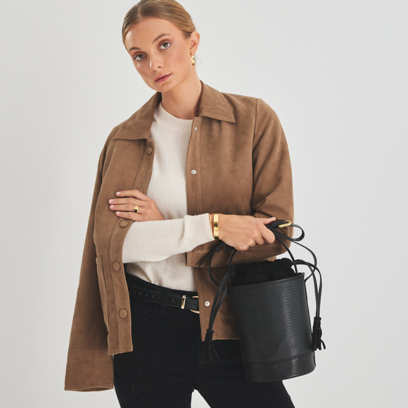 Bolsa bucket redonda pequena de couro em cor de serpente preta, vista frontal, segurada pelas mãos na alça - MONPIEL