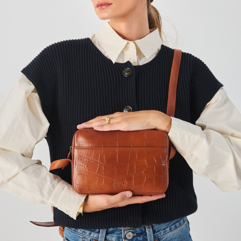 Bolsa de couro croco na cor couro com alça de bandoleira segurada com as mãos, vista frontal - MONPIEL