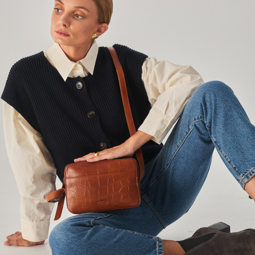 Bolsa de couro croco na cor couro com alça de bandoleira segurada com as mãos, modelo sentada no chão - MONPIEL