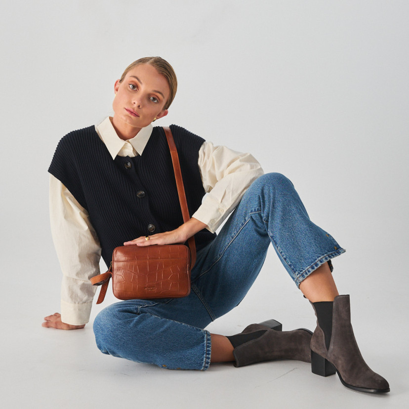 Bolsa de couro croco na cor couro com alça de bandoleira segurada com as mãos, modelo sentada no chão, plano geral - MONPIEL