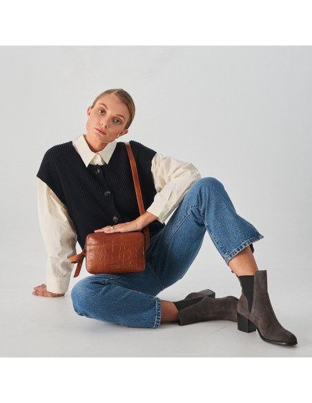 Crocodile leather crossbody bag in brown, with crossbody handle held with hands, model sitting on the floor, wide shot - MONPIEL