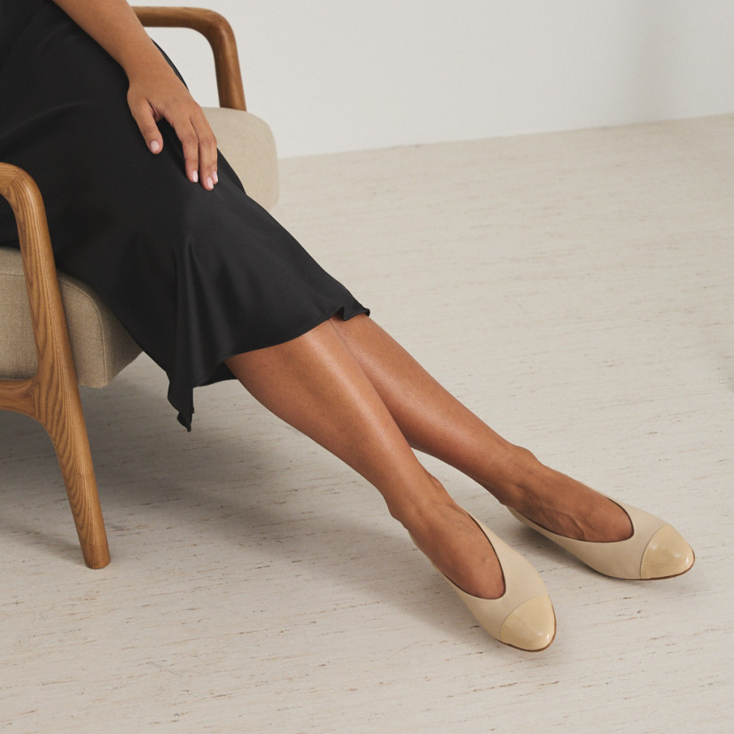 Bailarina de couro com salto e bico bicolor em bege/verniz, modelo sentada na cadeira com as pernas cruzadas para o la - MONPIEL