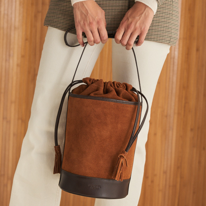 Bolsa redonda em forma de cubo de couro em camurça cor couro, vista frontal, fechada, segurada pelas mãos - MONPIEL
