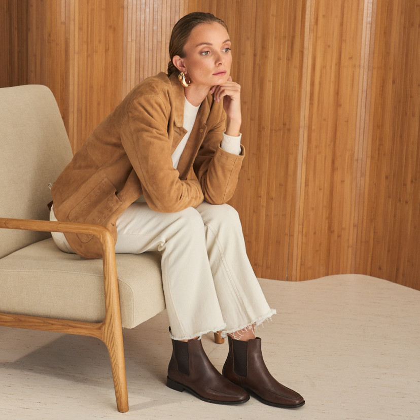 Luisa Chelsea ankle boot in brown leather with perspective view, wide shot of the model sitting sideways on the chair - MONPIEL