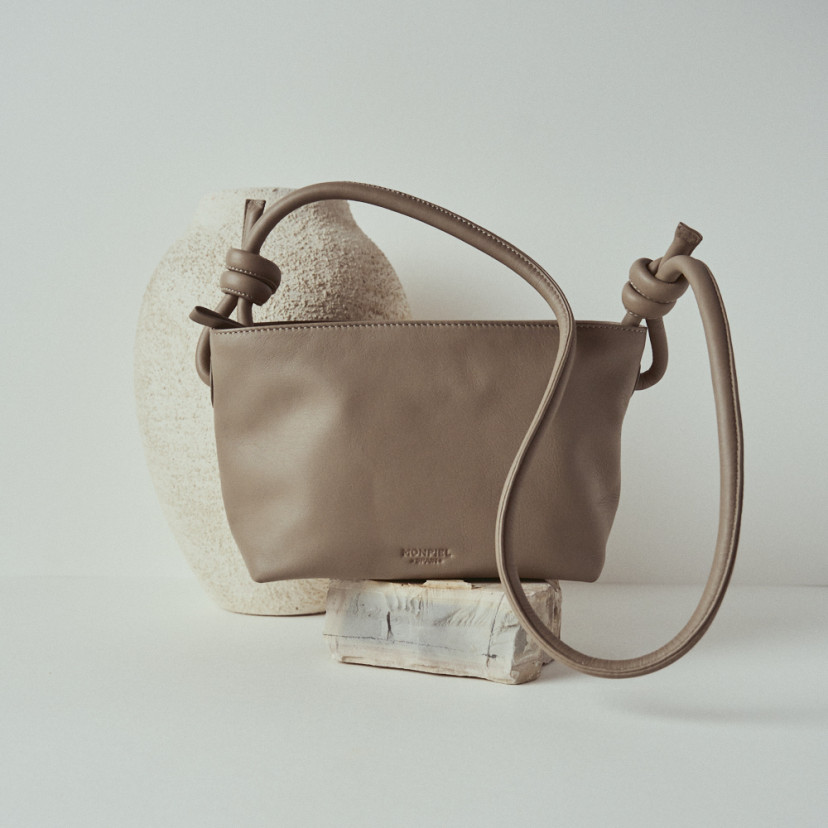 Bolsa pequena de couro com nós na cor taupe, vista frontal e detalhe da alça, apoiada sobre uma rocha - MONPIEL