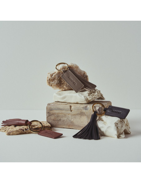 Porte-clés en cuir avec pompon noir, taupe et cuir, posé parmi des rochers - MONPIEL Porte-clés en cuir avec pompon noir, taupe et cuir, posé parmi des rochers - MONPIEL