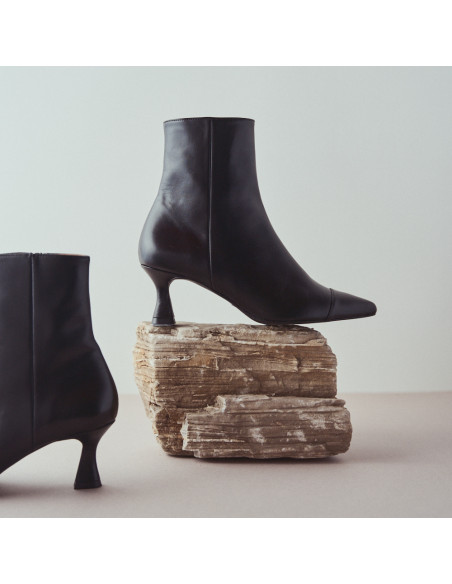 Black pointed-toe Jimena leather boot, profile view, resting on a rock, with the other boot beside it - MONPIEL Black pointed-toe Jimena leather boot, profile view, resting on a rock, with the other boot beside it - MONPIEL