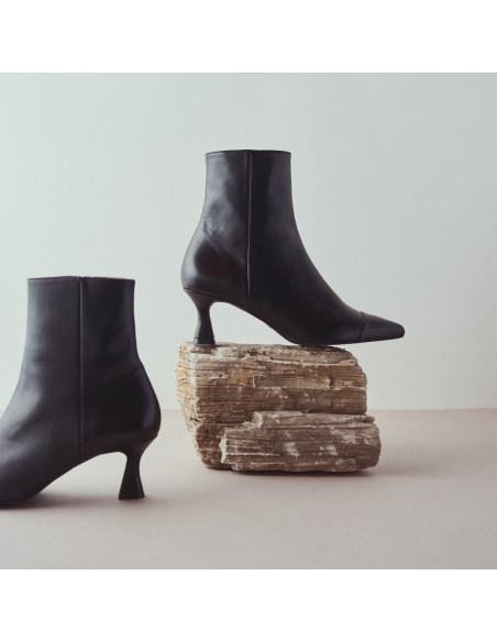 Black pointed-toe Jimena leather boot, profile view, resting on a rock, with the other boot beside it - MONPIEL Black pointed-toe Jimena leather boot, profile view, resting on a rock, with the other boot beside it - MONPIEL