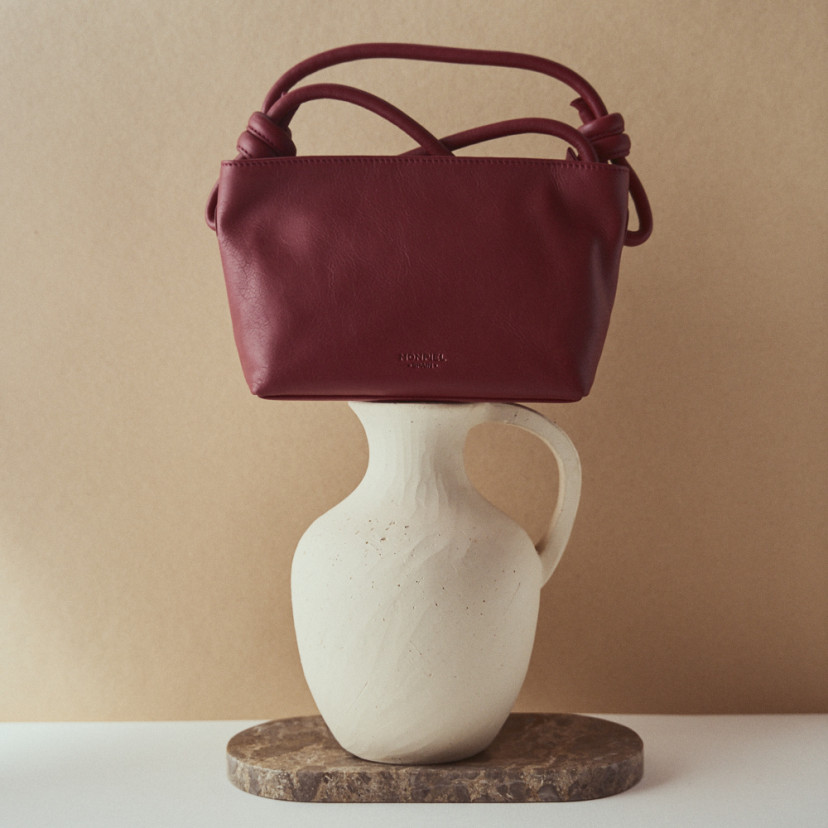 Bolsa pequena de couro com nós na cor bordeaux, vista frontal apoiada sobre um vaso - MONPIEL