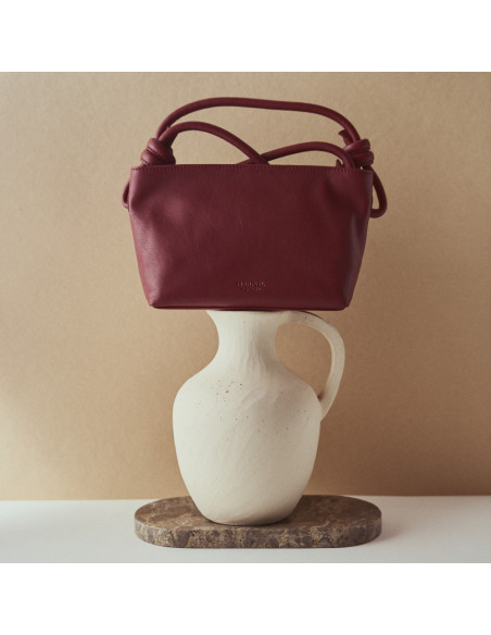 Kleine bordeauxfarbene Ledertasche mit Knoten, Frontalansicht, auf einem Vase abgestützt - MONPIEL Kleine bordeauxfarbene Ledertasche mit Knoten, Frontalansicht, auf einem Vase abgestützt - MONPIEL