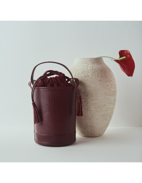 Piccola borsa a secchiello rotonda in pelle color bordeaux con effetto serpente, vista frontale, accanto a un vaso - MONPIEL