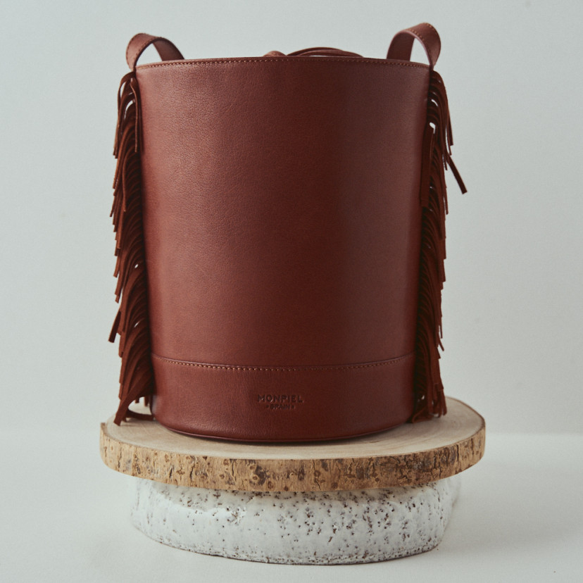 Bolsa de couro grande tipo bucket com franjas Roma na cor couro, close-up, vista frontal, apoiada em madeira - MONPIEL