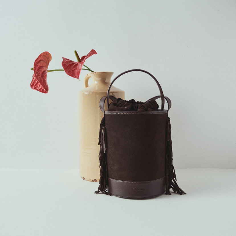 Bolsa pequena tipo bucket em camurça marrom Roma com franjas, vista frontal, em frente a um vaso com flor - MONPIEL